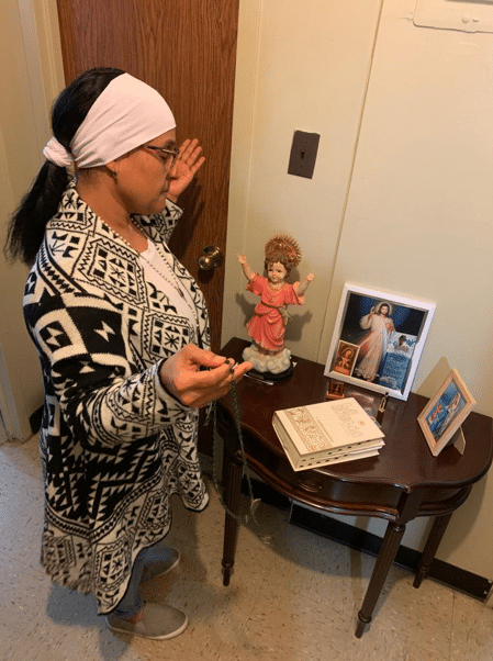 A woman stands by a small table  with hands raised, praying the Rosary.