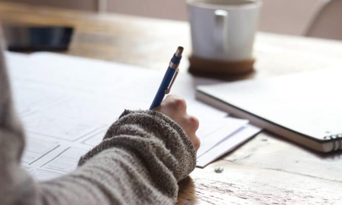 A student's arm and hand are visible as they do schoolwork at a table at home