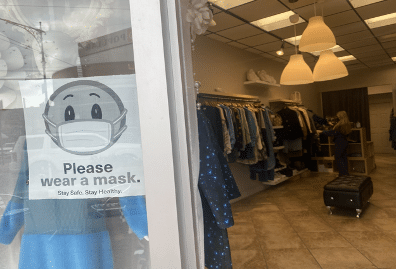A small clothing ship photographed from the doorway. Clothes are hanging on racks by the wall and a "Please were a mask" sign is seen in the window.
