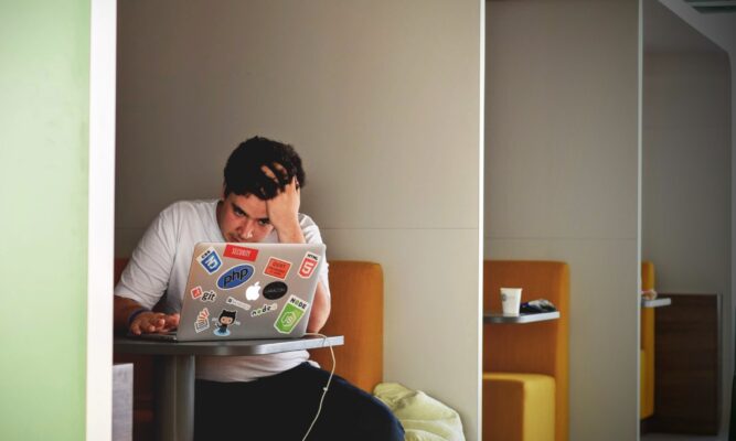 A student sits at a table, hand on his head, and stares at his laptop - looking stressed.