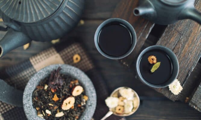 The angle is from up above looking down on two full cups of a dark liquid that looks like tea or black coffee. The cups are a deep teal. There are two matching teapots. There's also a bowl of loose tea that looks like potpourri.