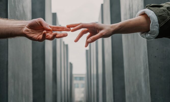 Two arms and hands reaching out – barely touching fingertips – with tall, uniformed, grey buildings in the background.