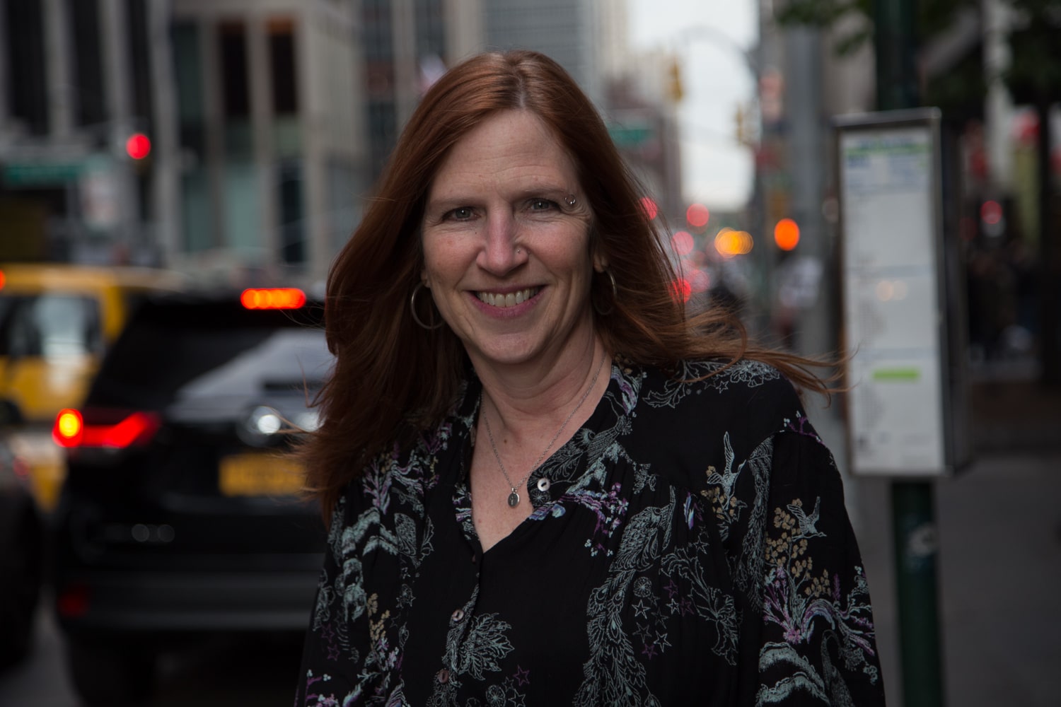 smiling woman with long red hair smiling at the camera. Busy city street behind her.