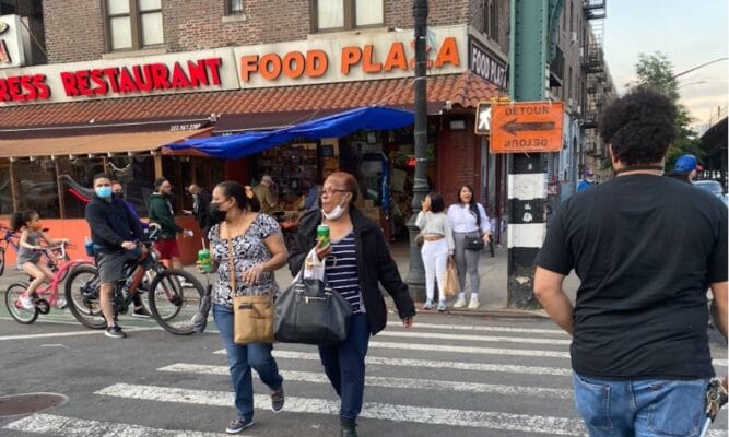 street view with people walking across the intersection. We also see an adult and kids on bikes. The main focus is a corner store and we see part of the name: "...RESS RESTAURANT" and the sign beside it says "FOOD PLAZA." This is the food plaza (bodega type store) mentioned in the article: Moronto’s Food Plaza on Dyckman Street in Upper Manhattan.