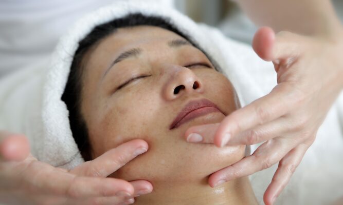 We see a woman's face (she looks Asian). Her head is wrapped in a towel. She's lying on her back with her eyes closed - peaceful. Two hands are massaging her face. She's receiving a facial treatment.
