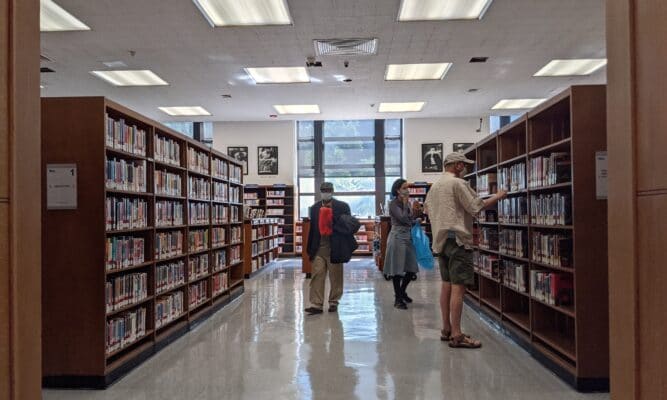Three people in masks, two that appear to be men and one woman (all in their 40s-50s) browse in a library aisle full of books. Fluorescent lights are on the ceiling. The floor is tile and shiny. We see windows at the far end. The aisle is wide.