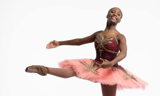 a young woman in a burgundy and pink leotard and tutu smiles and holds a dance pose with her right leg extended in front of her.